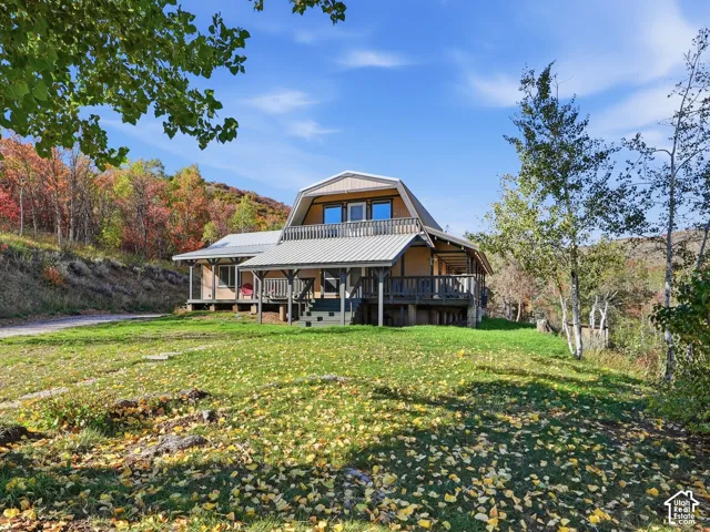 Gambrel-roof architectural style residence featuring a metal roof, wraparound wooden deck, upper-level balcony, and natural wooded surroundings