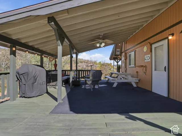 Covered exterior deck featuring a wood-plank floor, exposed beam ceiling, built-in ceiling fan, exterior wall-mounted light fixtures, and a protective railing