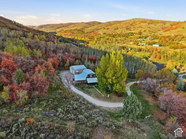 Multi-level residence featuring a gambrel roofline, attached metal workshop, gravel driveway, and surrounding mountainous terrain with dense deciduous vegetation
