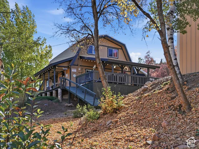 Gambrel-style exterior featuring vertical wood-finish siding, an expansive wraparound deck with integrated railing, and a ramp access point, situated on a sloped wooded lot