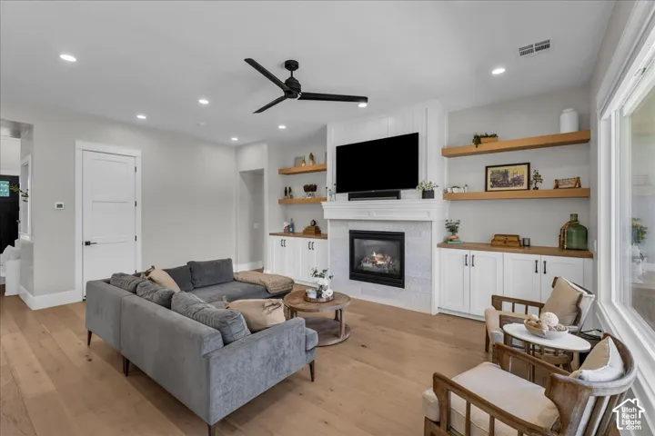Light wood-finish flooring, recessed ceiling lighting, a ceiling fan, a fireplace with a white shiplap surround, and built-in cabinetry with wood countertops and open shelving