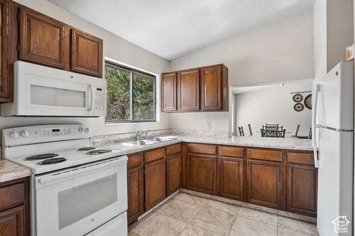Kitchen featuring wood-finish cabinetry, white appliances, a double basin sink, stone-look countertops, and tiled flooring