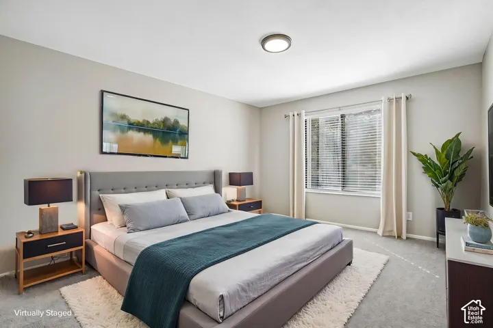 Master bedroom suite featuring a upholstered gray headboard, layered bedding in neutral tones, wood-finish nightstands with integrated lighting, large windows with cellular shades and grommet curtains providing natural light, and neutral carpeting through
