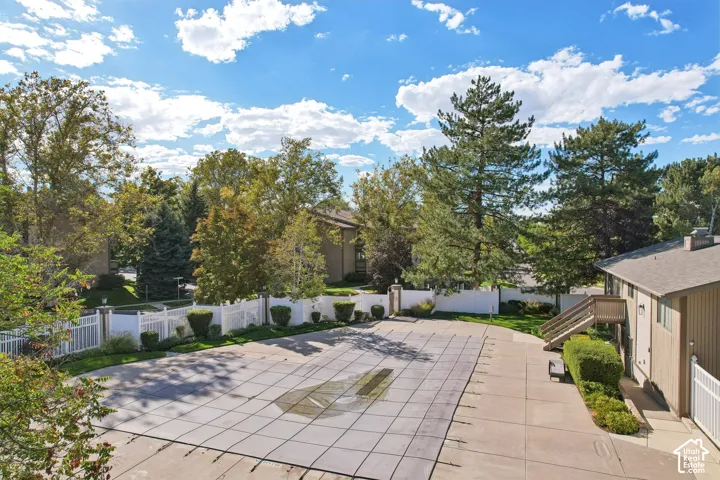 Covered pool with a surrounding paved deck, white fencing, and mature trees