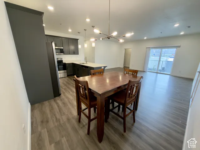 Open-concept living and dining area featuring wood-finish flooring throughout, a contemporary pendant light fixture, and seamless integration with a modern kitchen showcasing dark cabinetry and stainless steel appliances