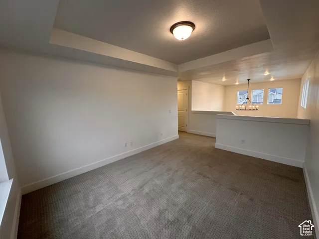 Tray ceiling with a flush mount light fixture, neutral carpeting, and a half-wall partition with a chandelier in the background