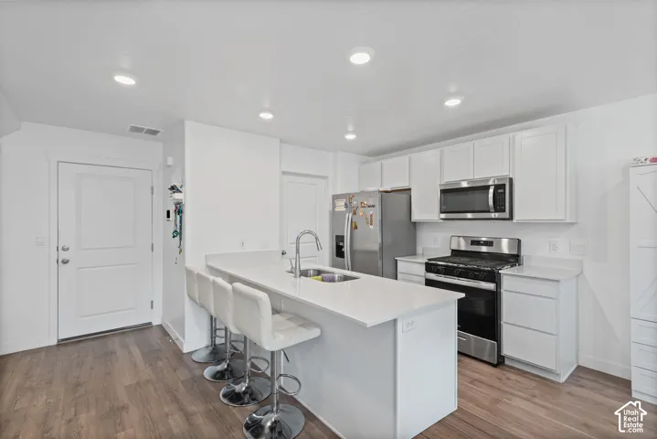 White kitchen cabinetry with stainless steel appliances, a center island with a sink, and wood-finish flooring