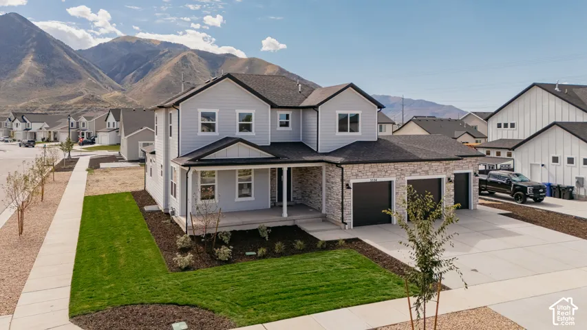 Modern two-story home featuring a clean architectural design with stone and siding exterior, expansive front-facing driveway with three-car garage, manicured lawn with fresh landscaping and young plantings, covered front porch entry, and mountain backdrop