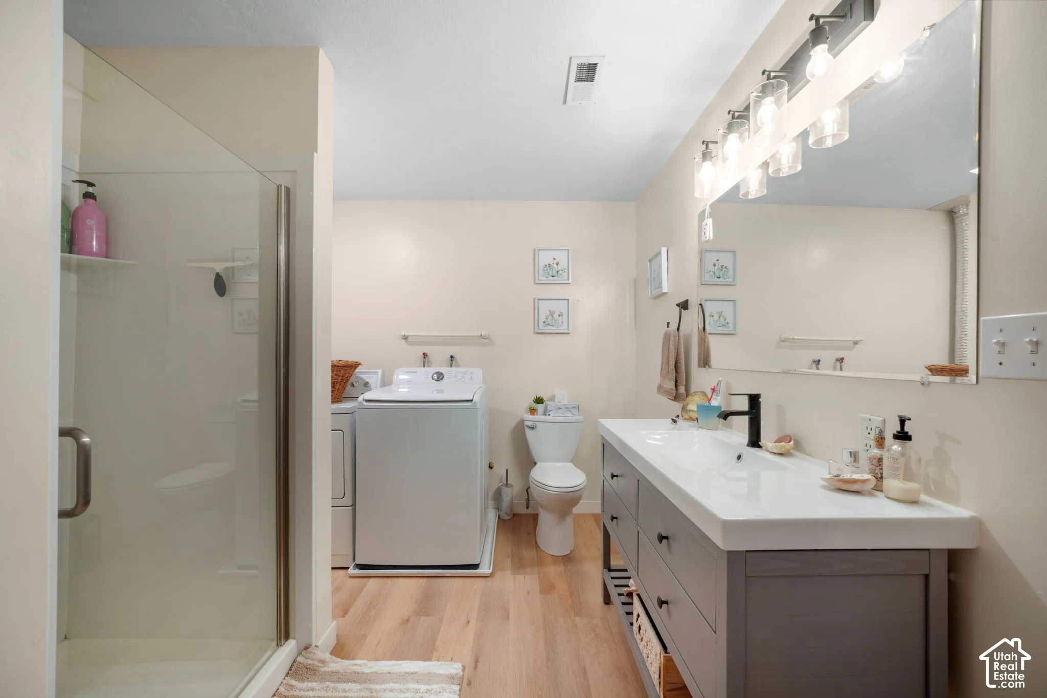 Shower with glass enclosure, single vanity with integrated sink, wood-finish flooring, and wall-mounted lighting