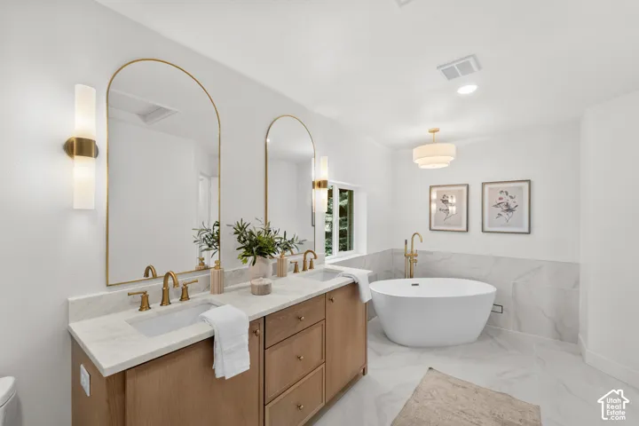 Dual arched mirrors with brass-finish frames, light wood-finish double vanity with stone countertop, freestanding soaking tub with brass-finish floor-mounted filler, and tile flooring