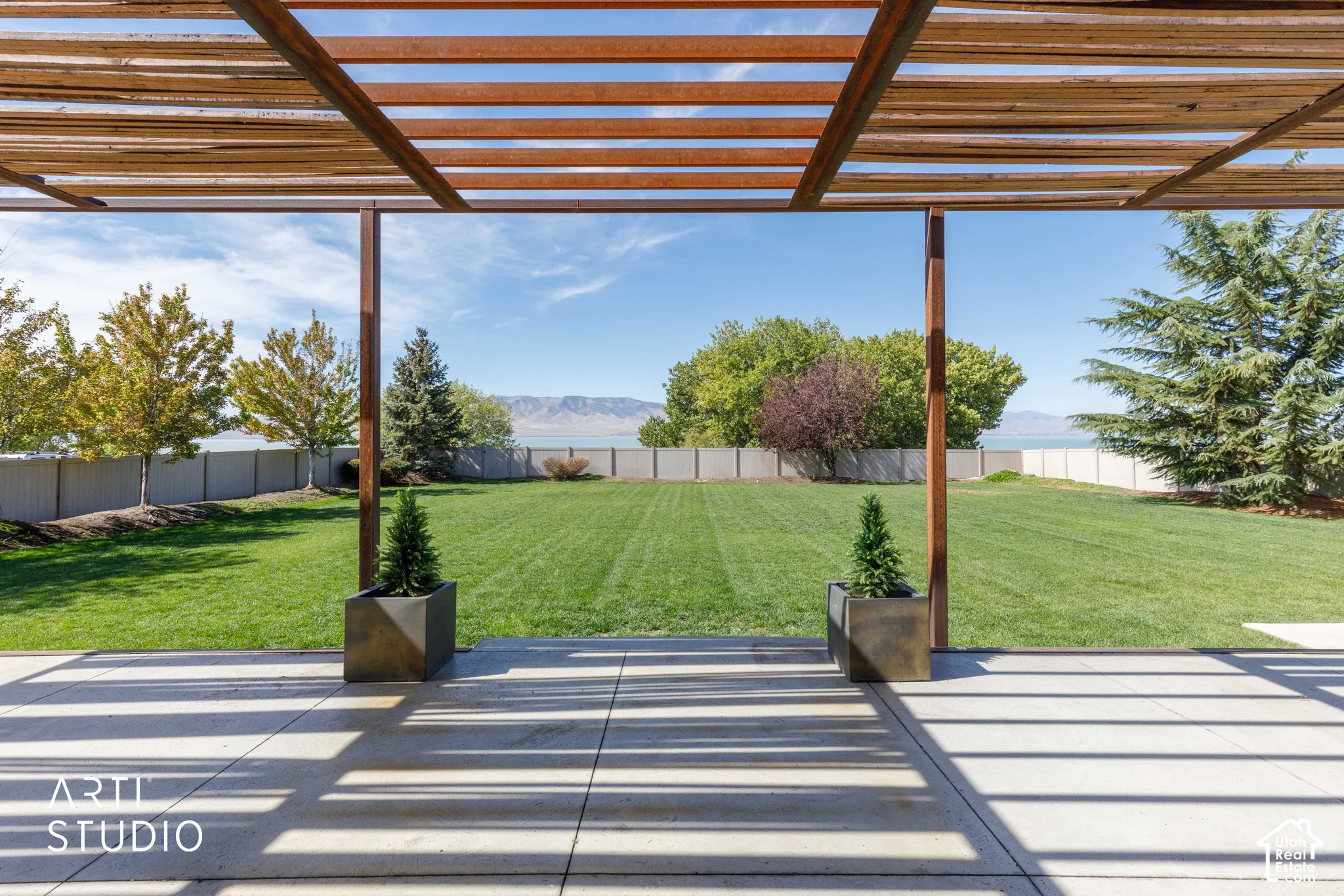 Expansive patio with concrete pavers and a wood-slat pergola, overlooking a large grass yard with mature trees and a distant body of water