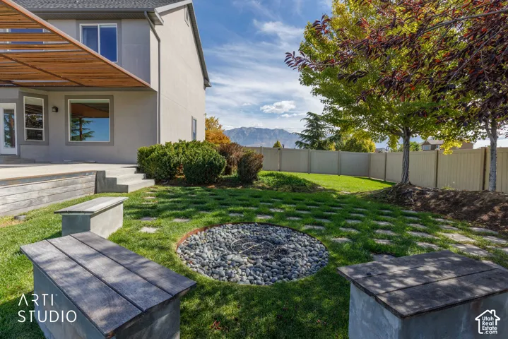 Expansive backyard featuring a built-in fire pit, stone paver pathway, concrete bench seating, mature trees, and a privacy fence
