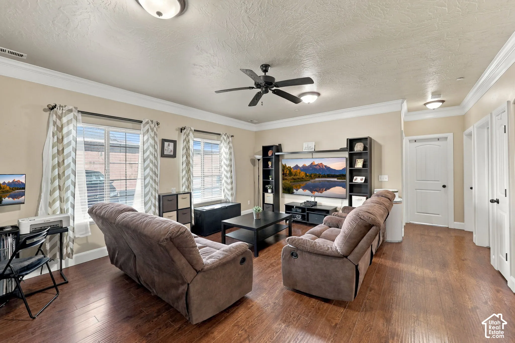 Spacious living area featuring wood-finish flooring, two windows with blinds, a ceiling fan with integrated lighting, and multiple flush-mount ceiling lights