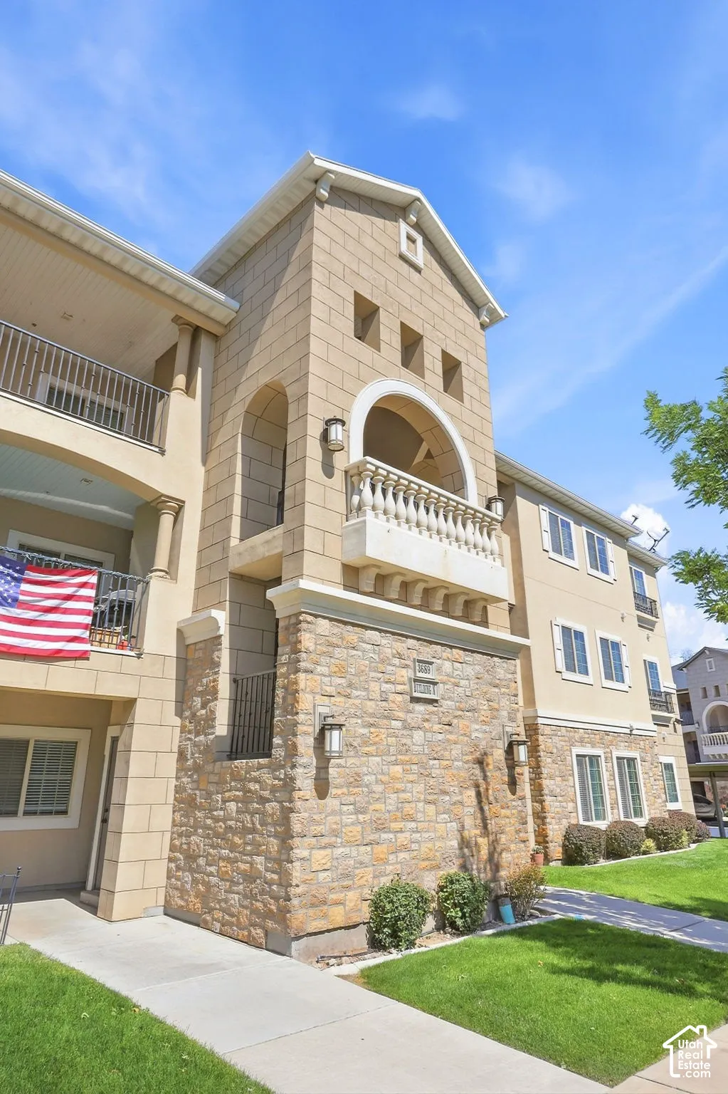 Stucco exterior with stone veneer accents, upper-level balcony with ornate balustrade, ground-level patio, and exterior wall sconces