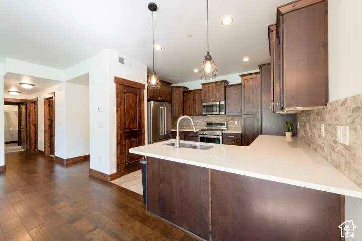 Kitchen featuring a peninsula island with an undermount sink, stainless steel appliances, dark wood-finish cabinetry, and a stone tile backsplash