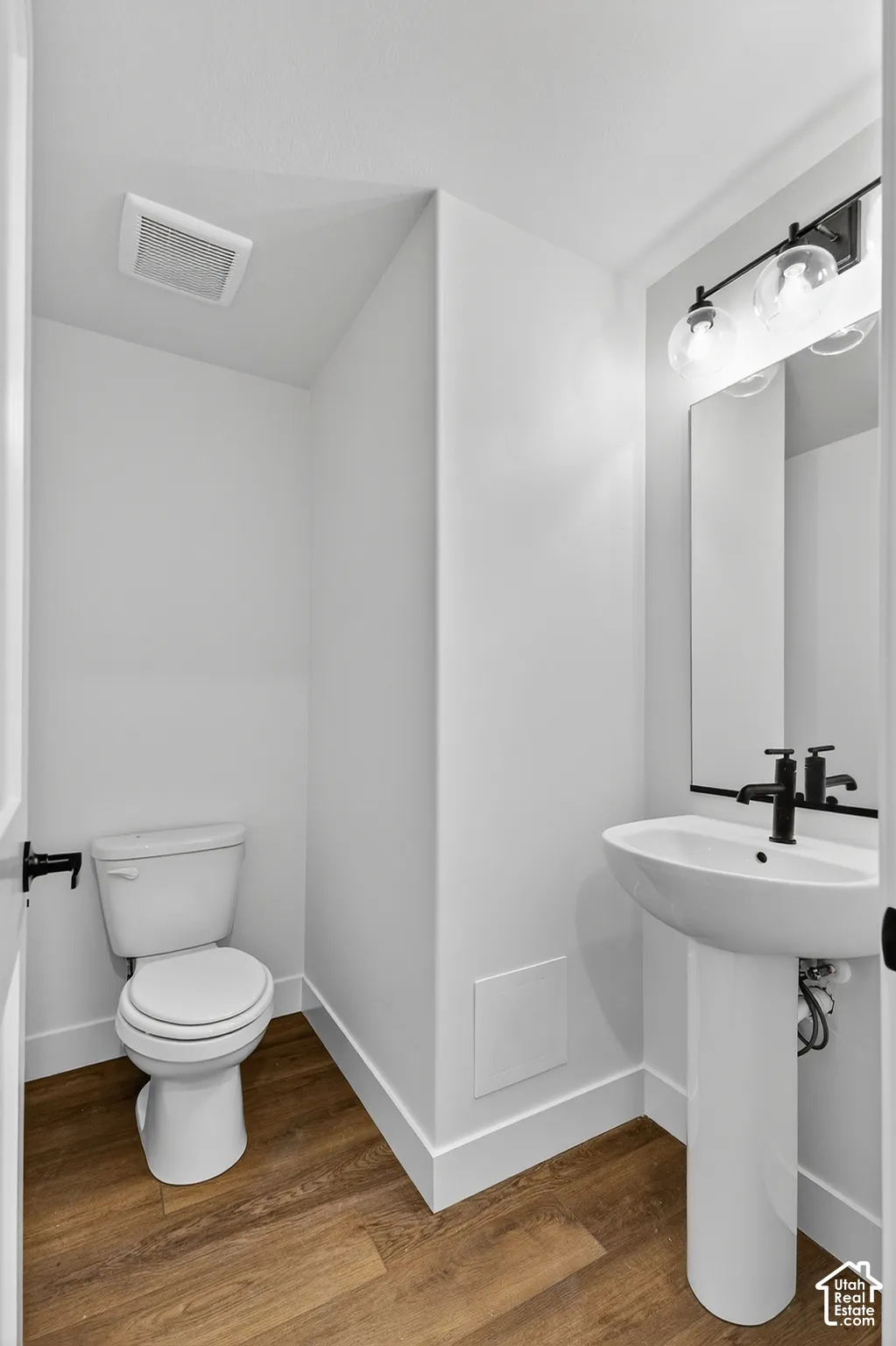 Pedestal sink with a matte black faucet, wood-finish flooring, and wall-mounted vanity lighting