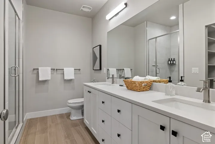 Dual vanity with integrated sinks, white cabinetry with dark hardware, a large wall mirror, a glass-enclosed shower, and wood-finish flooring