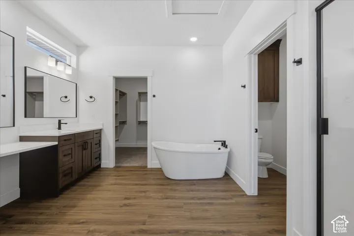 Dual vanity with white countertops, freestanding bathtub, wood-finish flooring, and a separate water closet