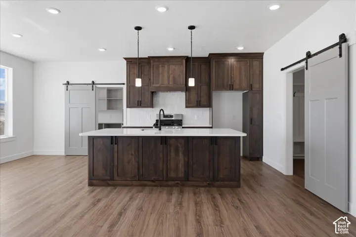 Contemporary kitchen featuring dark wood cabinetry, a large waterfall-edge island with seating, stainless steel appliances, white subway tile backsplash, pendant lighting, and wood-finish flooring throughout the open-concept space
