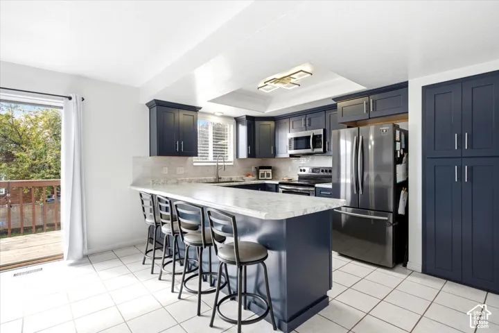 Kitchen featuring dark blue cabinetry, light tile flooring, a built-in breakfast bar, stainless steel appliances, and a sliding glass door to an outdoor deck