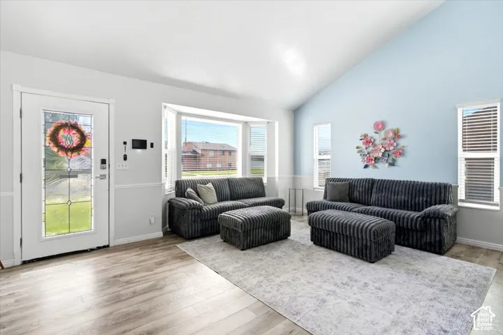 Vaulted ceiling, wood-finish flooring, bay window, white paneled entry door with decorative glass, and multiple windows with blinds