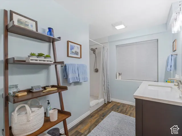 Bathroom featuring a dark wood-finish vanity with a white countertop, a shower with a white base, wood-finish flooring, a wall-mounted shelving unit, and an overhead light fixture