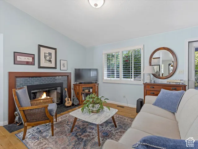 Living area with a fireplace featuring a tile surround, hardwood-finish flooring, plantation shutters, and a vaulted ceiling