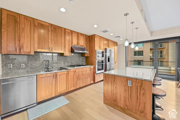 Kitchen featuring wood-finish cabinetry, stainless steel appliances, a granite backsplash, and a granite island with pendant lighting