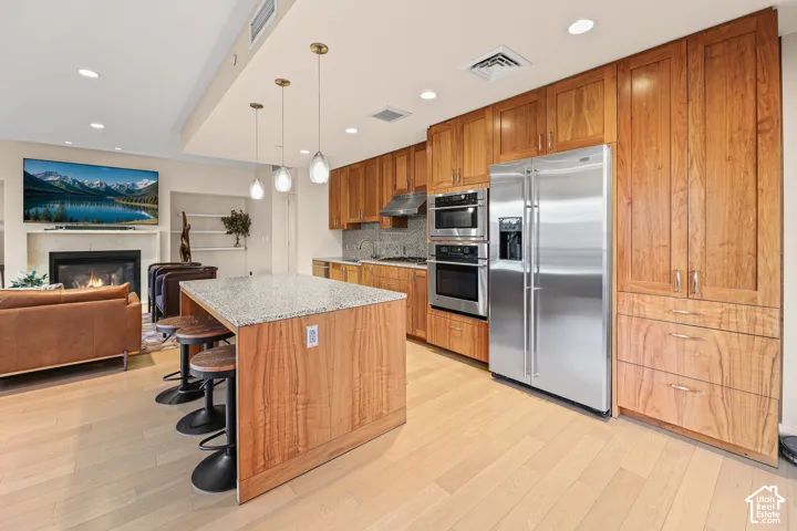 Open-concept living space with light-tone wood-finish flooring, a kitchen island with a stone countertop, wood-finish cabinetry, stainless steel appliances, and a fireplace with a stone mantel