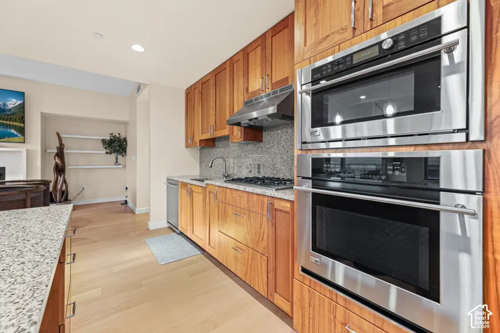 Chef's kitchen featuring warm wood cabinetry, stainless steel dual wall ovens with microwave insert, quartz countertops, tile backsplash, and wood-finish flooring throughout the open layout