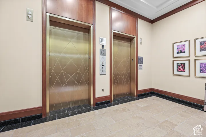 Two elevators with patterned brass doors, wood paneling surrounds, light-toned tiled flooring, and dark baseboard trim