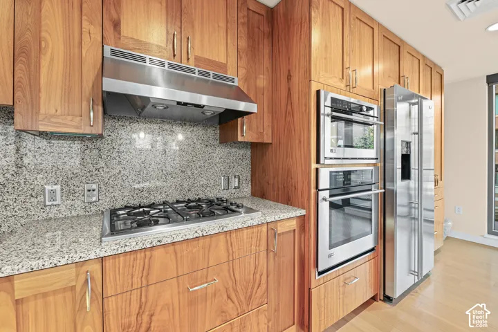 Wood cabinetry with stainless hardware, stainless steel gas range, stainless steel professional-grade hood, integrated stainless steel double ovens, and a stainless steel side-by-side refrigerator