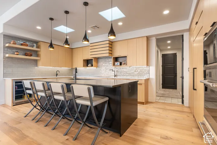 Kitchen featuring light wood-finish cabinetry, a large center island, integrated beverage refrigerator, built-in oven, and skylights