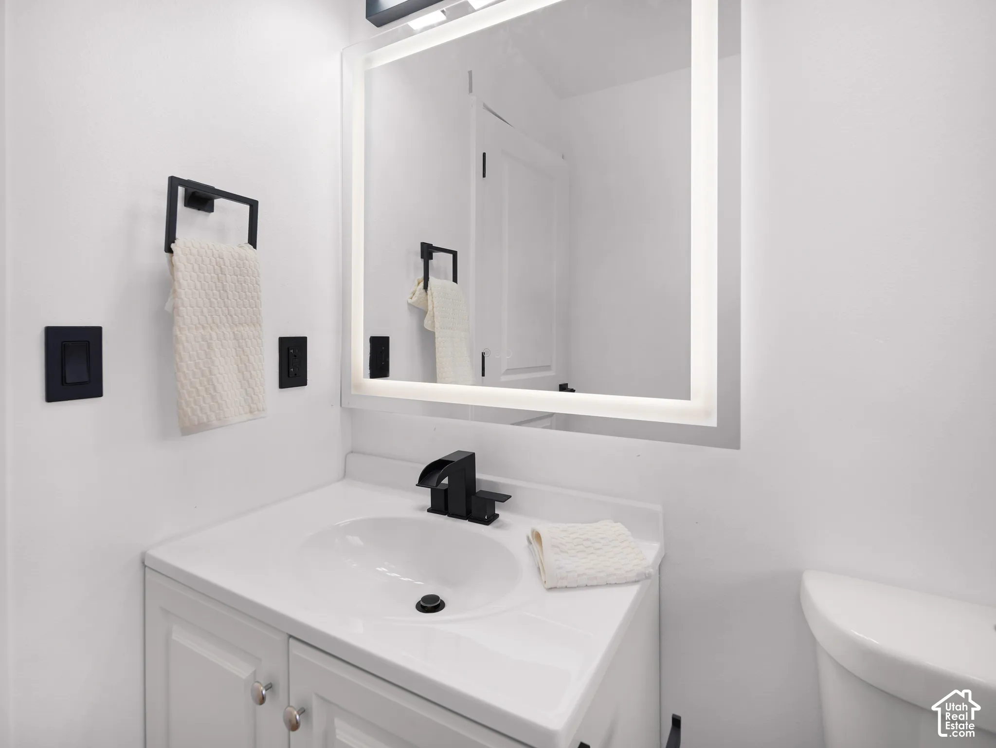 White vanity with a single basin sink, matte black waterfall faucet, and an illuminated mirror