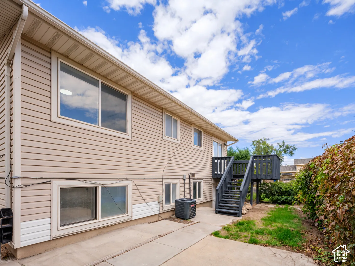 Beige siding exterior with multiple windows, concrete patio, and dark-painted deck with stairs