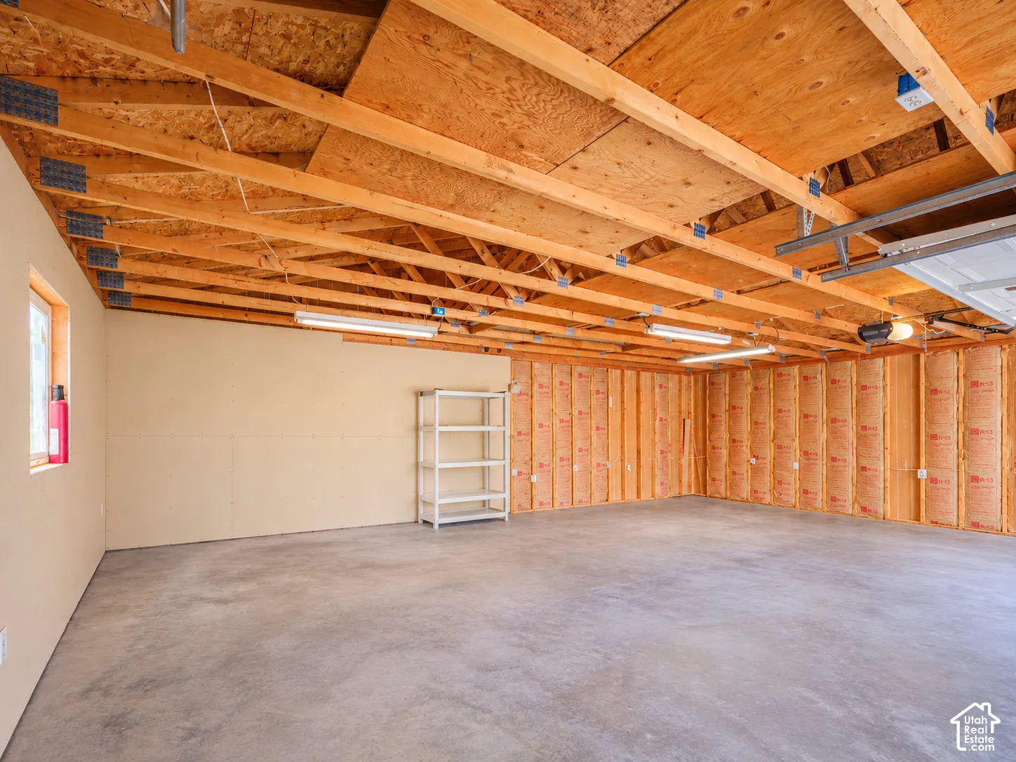 Exposed wood beam ceiling with plywood sheathing, concrete flooring, fluorescent lighting fixtures, and a garage door opener mechanism