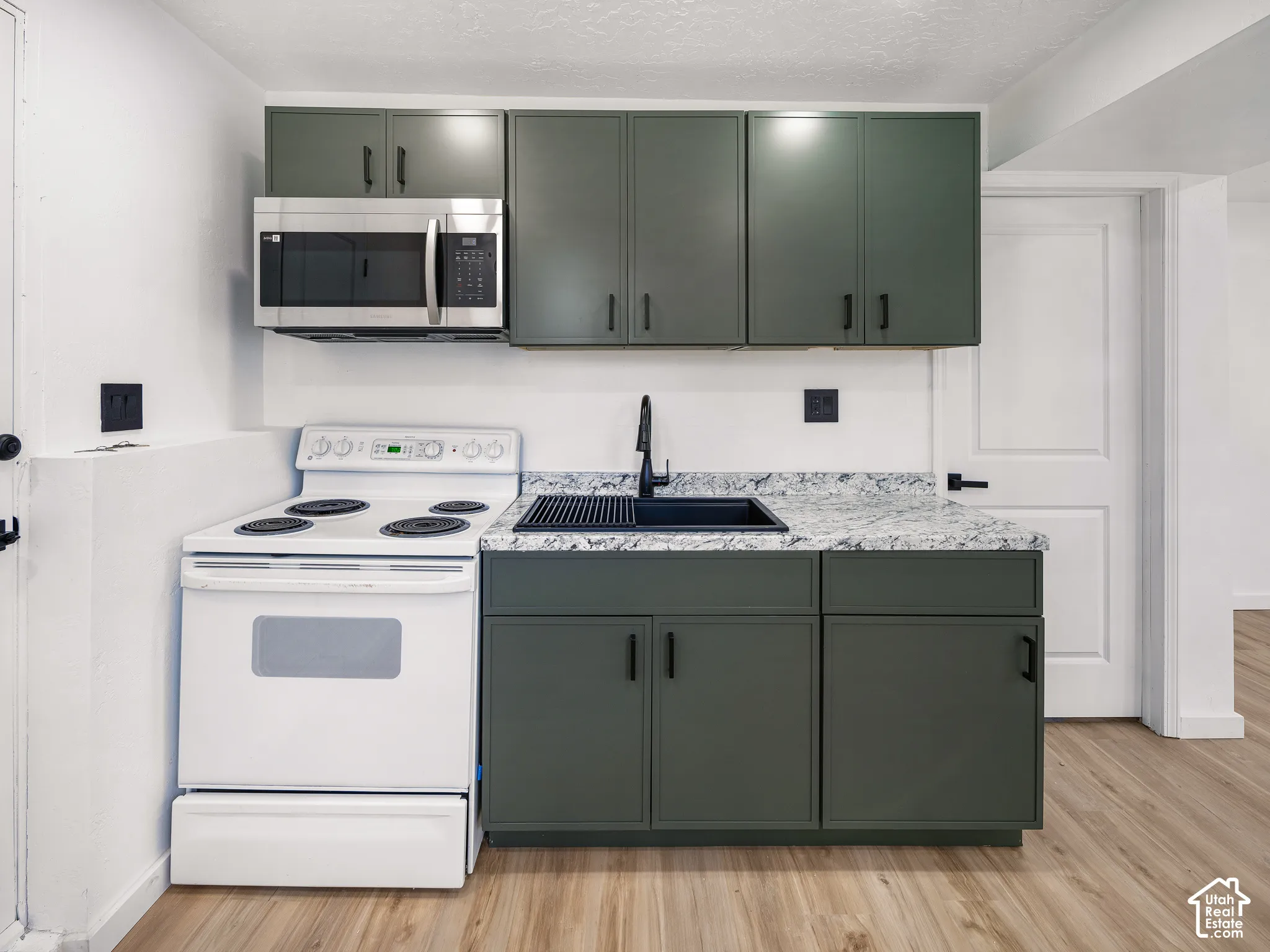 Kitchenette featuring olive green cabinetry, light-toned wood-finish flooring, stainless steel microwave, black sink, and speckled countertops