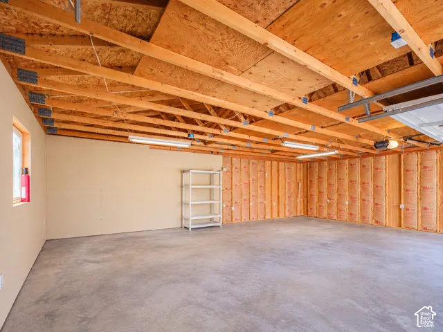 Exposed wood beam ceiling with plywood sheathing, concrete flooring, fluorescent lighting fixtures, and a garage door opener mechanism