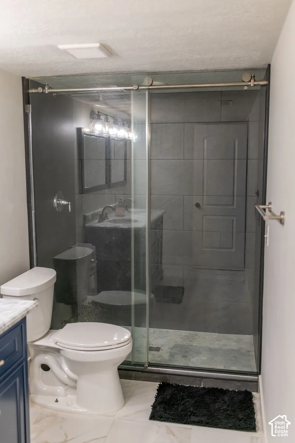 Glass-enclosed shower with sliding door, tiled shower surround, recessed shower pan, white toilet, and marble-finish tile flooring