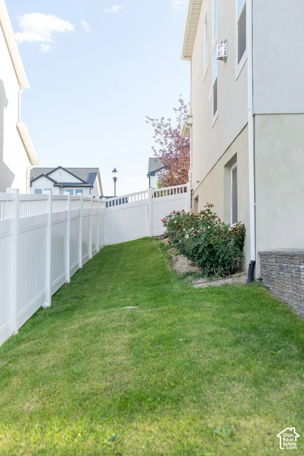Expansive side yard featuring a manicured lawn, white privacy fencing, mature shrubbery, and stone veneer foundation detailing