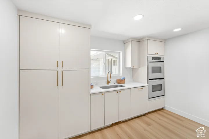 Full-height cabinetry, under-mount sink with a gold-finish faucet, white countertops, built-in double ovens, and wood-finish flooring