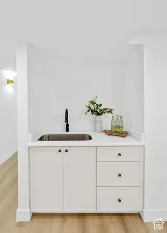 Wet bar featuring a stainless steel sink, solid surface countertop, white cabinetry, matte black faucet, and wood-finish flooring