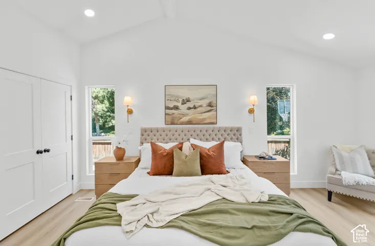 Vaulted ceiling with recessed lighting, light wood-finish flooring, two large windows, white double closet doors, and wall-mounted sconces