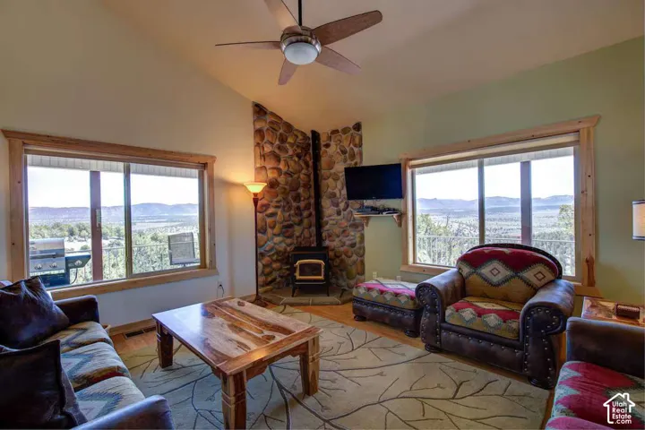 Vaulted ceiling with a ceiling fan, stone-clad fireplace with a wood-burning stove, large windows with natural wood trim, and wood-finish flooring