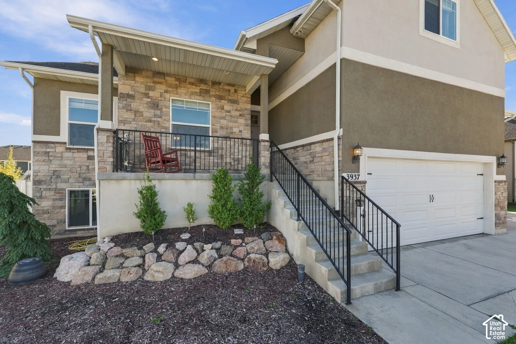 Multi-level exterior featuring a stone veneer facade, stucco siding, elevated covered porch with black metal railing, and concrete driveway leading to an attached two-car garage
