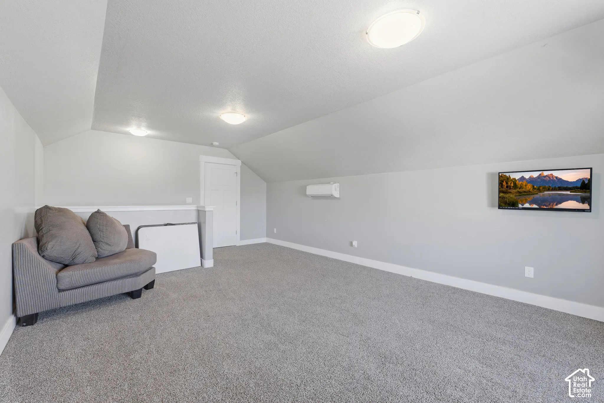 Vaulted ceiling, wall-mounted ductless climate control unit, carpeted flooring, and flush-mount ceiling lighting
