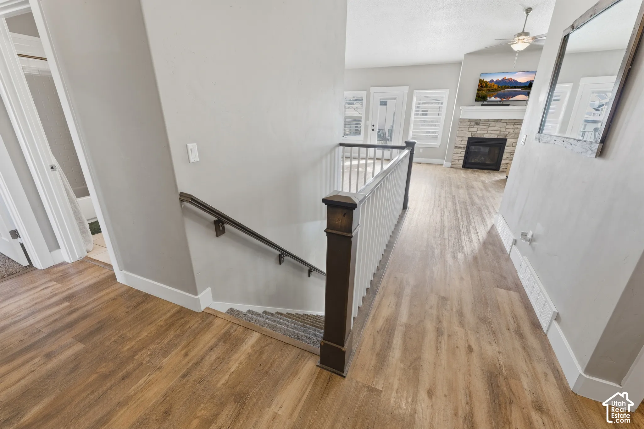 Wood-finish flooring throughout, staircase featuring a dark-stained wood newel post and balustrade, and a stone-surround fireplace with a mantel