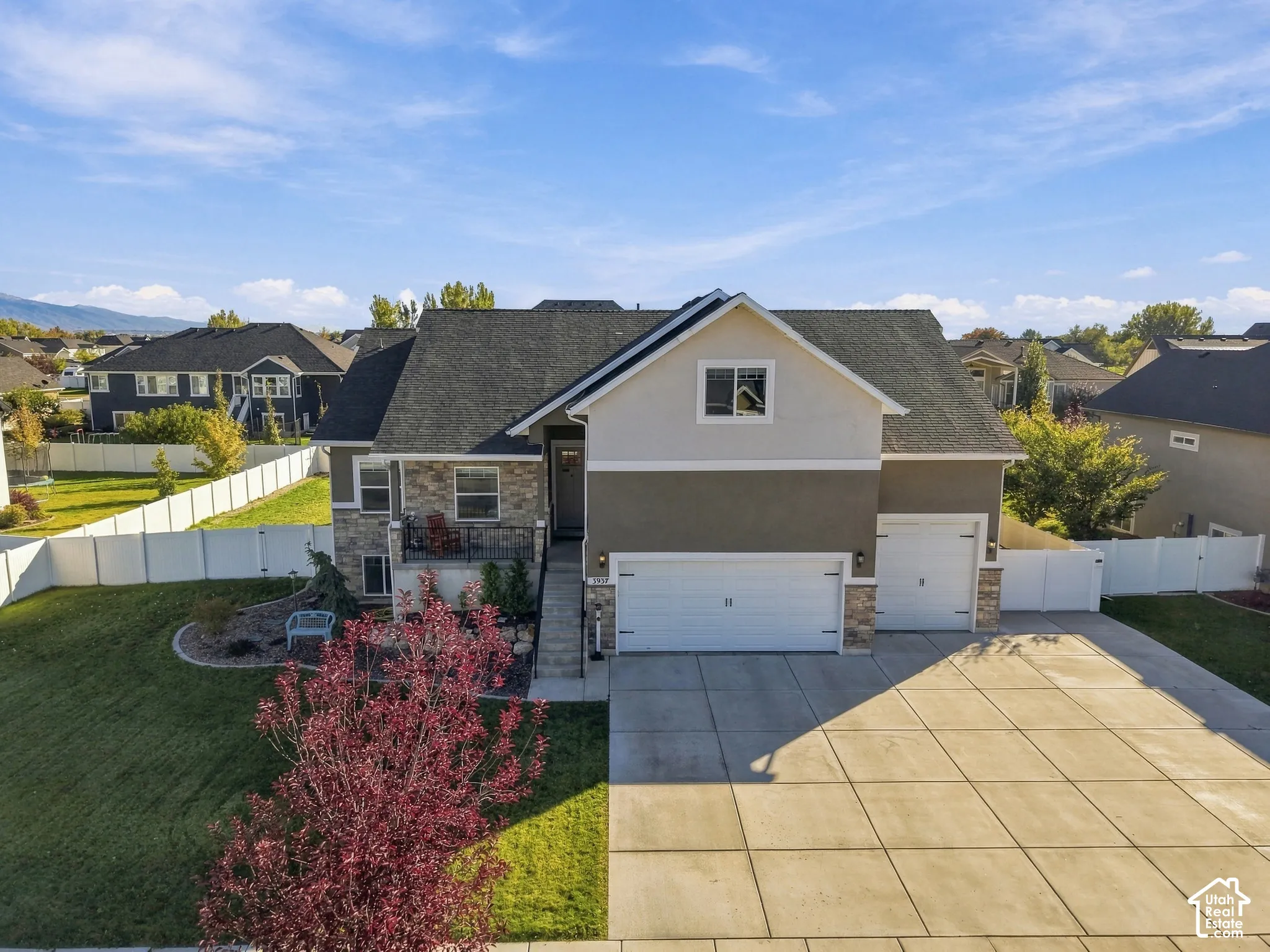 Two-story exterior featuring a mix of stucco and stone veneer siding, a three-car garage with concrete driveway, and white perimeter vinyl fencing