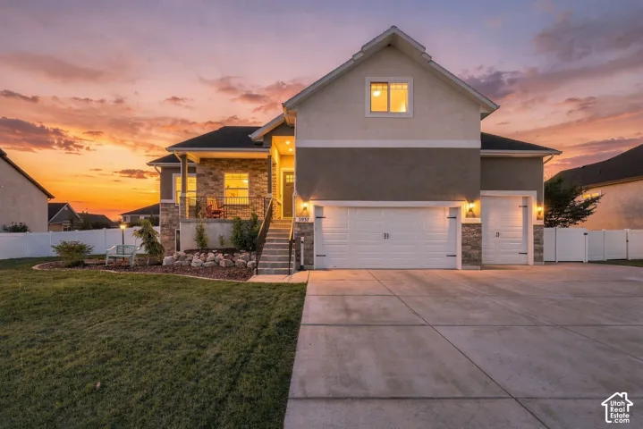 Edited Twilight Photo. Two-story residence featuring a mixed-material exterior of stone veneer and stucco, a three-car garage, concrete driveway, and an elevated front porch