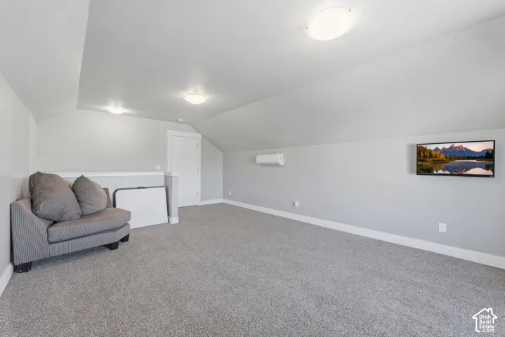 Vaulted ceiling, wall-mounted ductless climate control unit, carpeted flooring, and flush-mount ceiling lighting